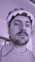 a selfie of a white person, wearing either headphones or a helmet, making an odd expression. the photo is edited to be colorized into a monochrome purple-ish color palette.